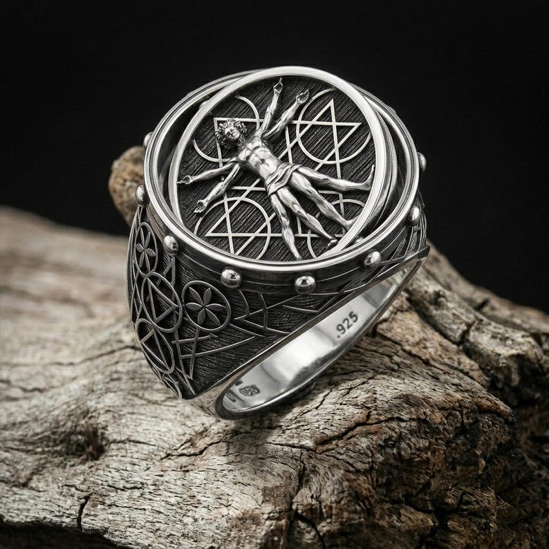 May include: A silver ring featuring a detailed depiction of the Vitruvian Man within a circular frame. The ring has intricate geometric patterns and the inscription ".925" on the band, indicating its silver composition. The ring is displayed on a piece of wood.