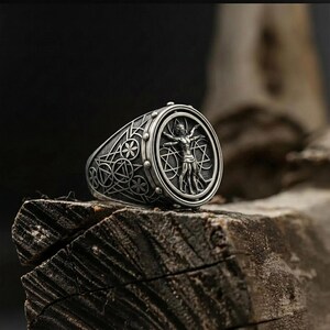 May include: A silver ring featuring a detailed depiction of the Vitruvian Man within a circular frame. The ring's band is adorned with geometric patterns and floral motifs. The ring is displayed on a piece of weathered wood.