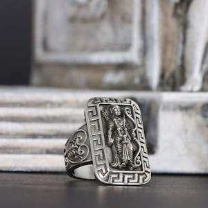 Może przedstawiać: Srebrny pierścionek z prostokątną tarczą z detalicznym reliefem przedstawiającym postać trzymającą łuk i strzałę. Pierścionek ma ozdobną obwódkę i szczegółowy pas z wirującym wzorem. Pierścionek jest na rozmytym tle.