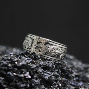 Pu&ograve; includere: Un anello d'argento con il numero 13 al centro, circondato da motivi floreali e a foglie. L'anello &egrave; posto su uno sfondo scuro e strutturato, che mette in risalto i dettagli intricati del design. Un gioiello.