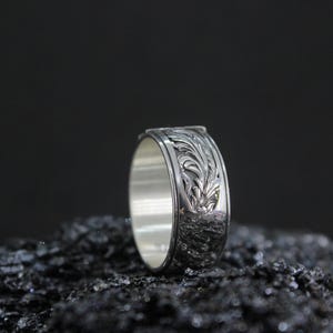 Pu&ograve; includere: Un anello d'argento con un motivo floreale inciso. L'anello &egrave; largo e ha un interno lucido. L'anello &egrave; esposto su una superficie scura e strutturata su uno sfondo nero. Il design dell'anello &egrave; dettagliato e intricato.