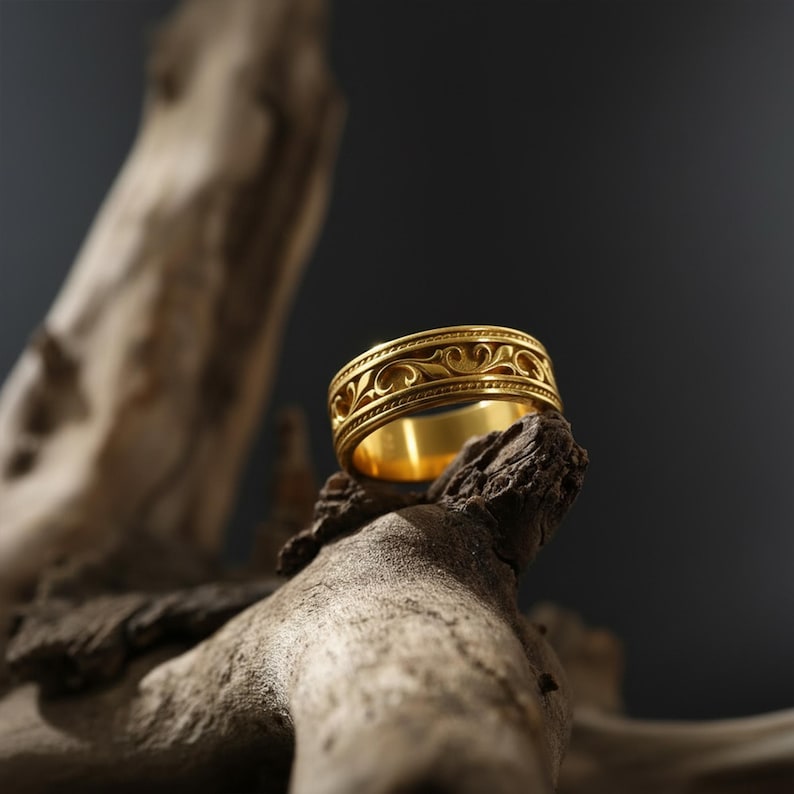 Puede incluir: Un anillo dorado con un intrincado patr&oacute;n floral repetitivo. El anillo se exhibe sobre un trozo de madera envejecida contra un fondo oscuro. El dise&ntilde;o del anillo presenta una serie de l&iacute;neas curvas y espirales, creando una apariencia detallada y ornamentada.