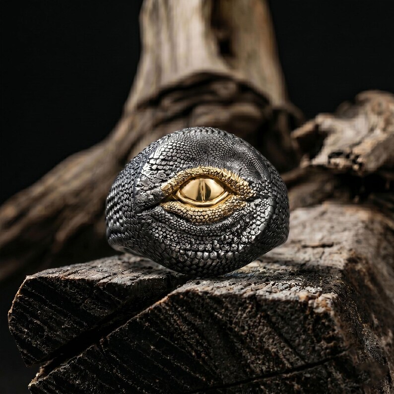 Puede incluir: Un anillo con una superficie oscura y texturizada de escamas de reptil en acabado gris plomo. El centro del anillo muestra una forma de ojo dorada, lisa y prominente, con p&aacute;rpados dorados detallados y texturizados alrededor del iris.