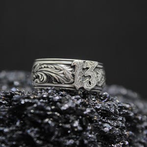 Pu&ograve; includere: Un anello d'argento con un ornato motivo floreale e il numero 13 in un carattere stilizzato. L'anello &egrave; posto su uno sfondo scuro e strutturato, che ne evidenzia gli intricati dettagli.