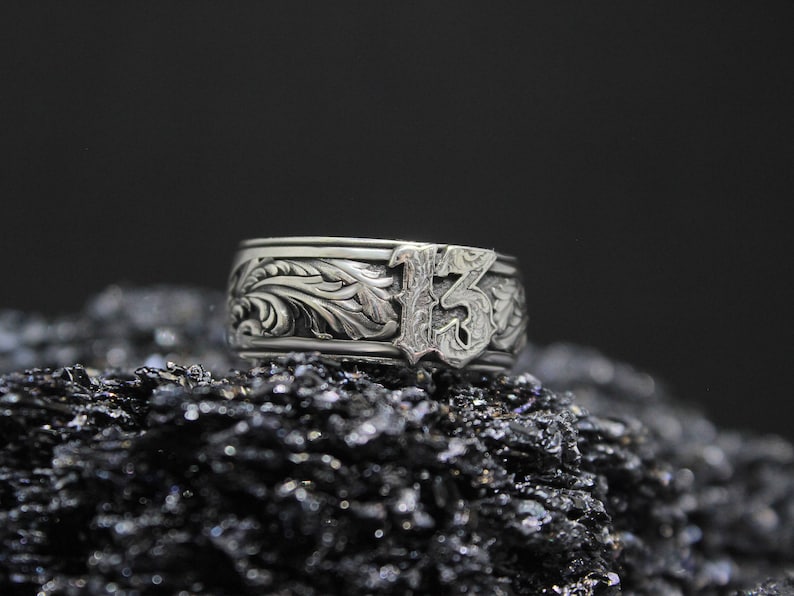 Pu&ograve; includere: Un anello d'argento con un ornato motivo floreale e il numero 13 in un carattere stilizzato. L'anello &egrave; posto su uno sfondo scuro e strutturato, che ne evidenzia gli intricati dettagli.