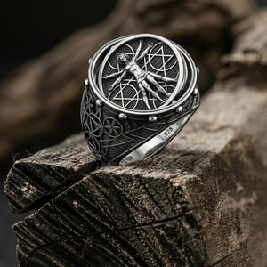 May include: A detailed silver ring featuring a central design of a human figure within a circle, surrounded by geometric shapes. The ring has a textured band and the marking ".925". The ring is displayed on a rough wooden surface.