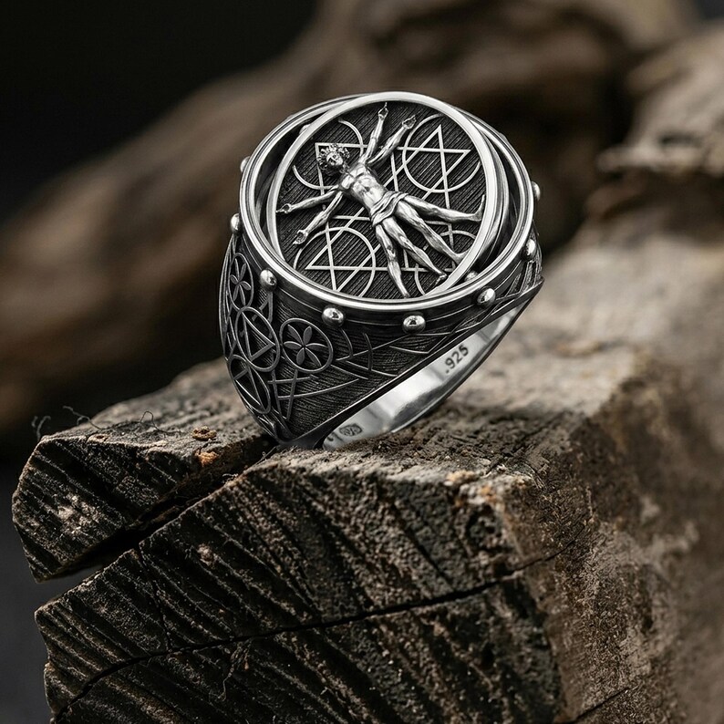 May include: A detailed silver ring featuring a central design of a human figure within a circle, surrounded by geometric shapes. The ring has a textured band and the marking ".925". The ring is displayed on a rough wooden surface.