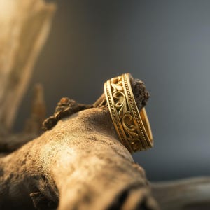 Puede incluir: Un anillo de oro con un patr&oacute;n ornamental y ondulado. El anillo se muestra sobre un trozo de madera envejecida, con un fondo gris oscuro. El dise&ntilde;o del anillo presenta un motivo floral repetido.