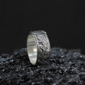 Pu&ograve; includere: Un anello d'argento con un intricato motivo floreale. L'anello &egrave; presentato su uno sfondo scuro, evidenziando il design dettagliato. La fascia presenta un motivo ripetuto di foglie e viti.