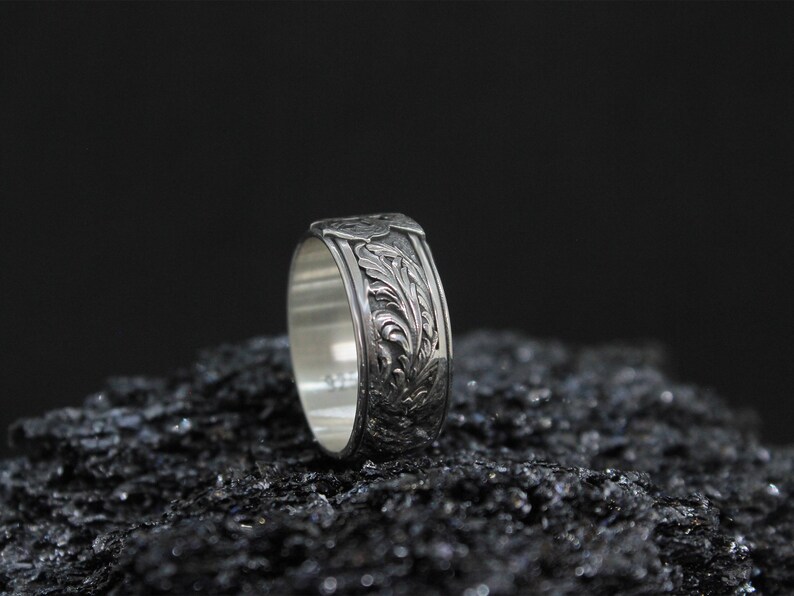 Pu&ograve; includere: Un anello d'argento con un intricato motivo floreale. L'anello &egrave; presentato su uno sfondo scuro, evidenziando il design dettagliato. La fascia presenta un motivo ripetuto di foglie e viti.