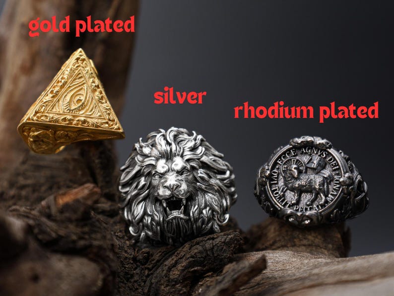May include: Three rings: a gold-plated pyramid, a silver lion head, and a rhodium-plated ring with a lamb design. The rings are displayed on a dark, textured surface, with the words "gold plated", "silver", and "rhodium plated" in red.