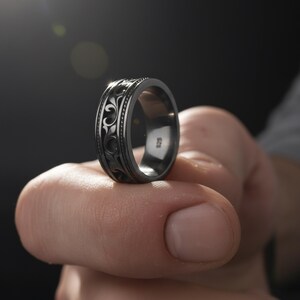 Puede incluir: Un anillo gris oscuro con un patr&oacute;n floral ornamentado y repetitivo. El anillo se sostiene, mostrando el intrincado dise&ntilde;o y el acabado pulido. El anillo es de estilo banda, con el n&uacute;mero "925" visible en el interior.