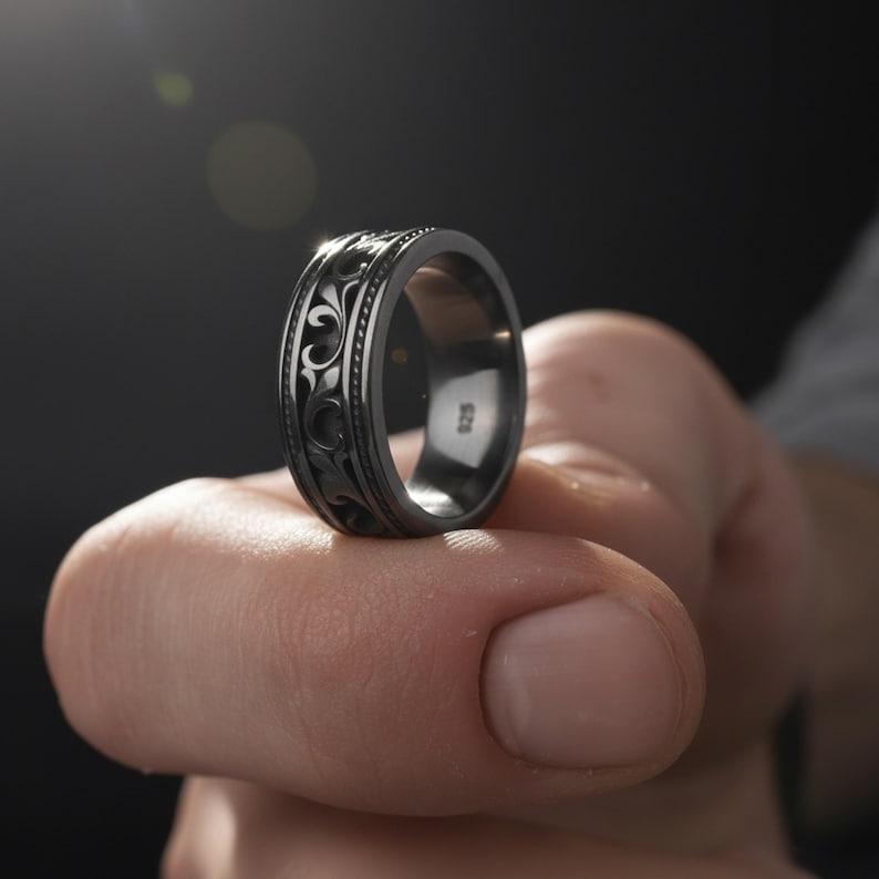Puede incluir: Un anillo gris oscuro con un patr&oacute;n floral ornamentado y repetitivo. El anillo se sostiene, mostrando el intrincado dise&ntilde;o y el acabado pulido. El anillo es de estilo banda, con el n&uacute;mero "925" visible en el interior.