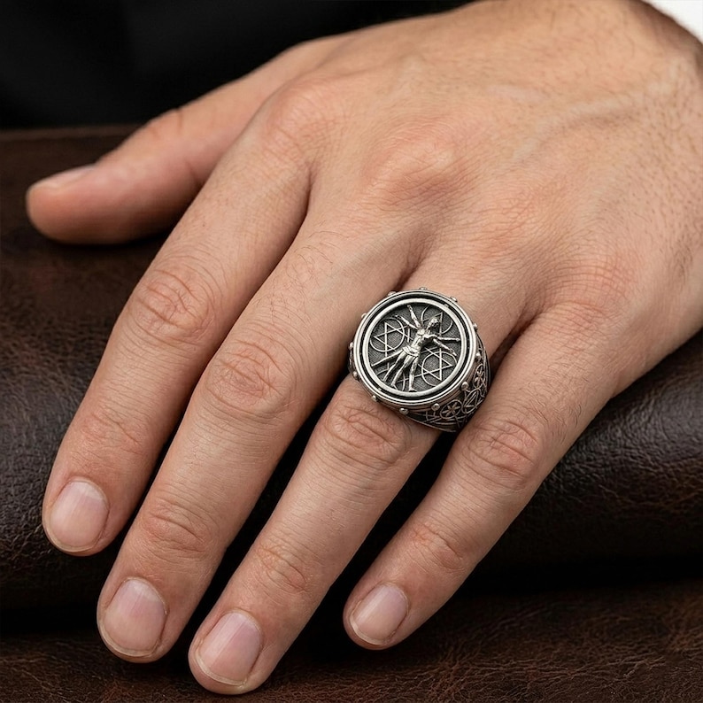 May include: A silver-toned ring featuring a detailed circular design with a central figure and surrounding geometric shapes. The ring is worn on a finger, showcasing its intricate craftsmanship and metallic sheen.