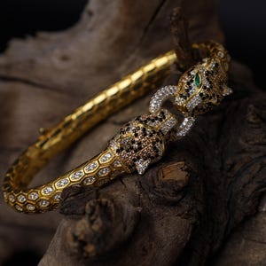 Könnte beinhalten: Goldfarbenes Armband mit Leopardenmuster. Das Armband hat ein strukturiertes Band und ist mit schwarzen, weißen und braunen Edelsteinen verziert. Die Augen des Leoparden sind grün. Das Armband wird auf einem Stück dunklem Holz präsentiert.