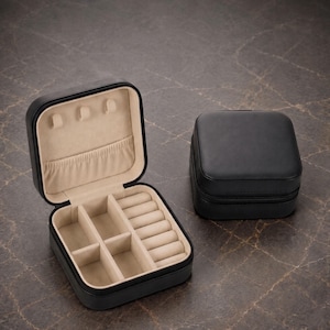 May include: A square black jewelry box with a zipper closure, shown both open and closed. The open box features a beige interior with three necklace hooks, a small elastic pocket, four open storage compartments, and six ring rolls for organization.