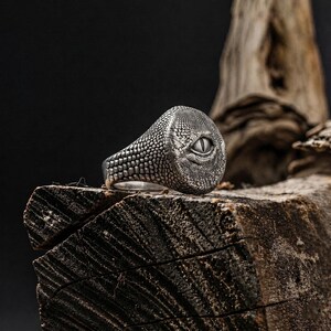 Puede incluir: Un anillo de plata con un patr&oacute;n texturizado de escamas de reptil en la banda y la cara. El centro del anillo muestra un ojo detallado en forma de almendra con una pupila vertical, elaborado con un acabado oscuro oxidado para resaltar.