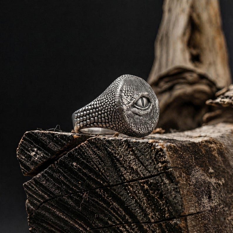 Puede incluir: Un anillo de plata con un patr&oacute;n texturizado de escamas de reptil en la banda y la cara. El centro del anillo muestra un ojo detallado en forma de almendra con una pupila vertical, elaborado con un acabado oscuro oxidado para resaltar.