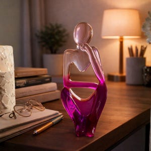 May include: A decorative glass sculpture of a person in a thinking pose. The sculpture is clear at the top, transitioning to a vibrant magenta at the base. It sits on a wooden surface, with a lamp and books in the background.