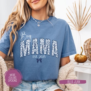 May include: A blue jean t-shirt with the words "Boy MAMA" in a floral design. The text "EST. 2025" and a bow are below the word "MAMA". The shirt has short sleeves and a round neckline.