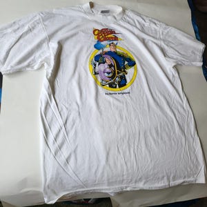 Puede incluir: Camiseta blanca con un gráfico de Captain Star, un personaje de cómic de Bernie Wrightson. El gráfico presenta a un hombre con un uniforme azul con un gran ojo en el pecho.