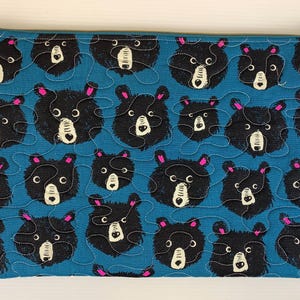 Puede incluir: Bolsa rectangular acolchada con fondo turquesa y un patrón repetido de caras de osos negros. Los osos tienen hocicos blancos, orejas rosas y están delineados con costuras blancas. Cremallera dorada y tirador turquesa y rosa.