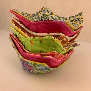 May include: A stack of patterned fabric bowl cozies. The cozies display a variety of designs, including floral and fruit patterns, and solid colours. The cozies are made of fabric and are designed to hold bowls of food. The colours include pink, green, yellow, and blue.