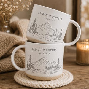 May include: Two off-white ceramic mugs stacked vertically, each featuring a gray line-art illustration of a mountain range, pine trees, and a river. The text "JAMES ♥ SOPHIA" and the date "04.08.2026" are printed in gray above the landscape.