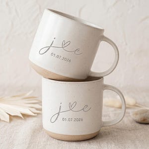 May include: Two speckled off-white ceramic mugs stacked vertically. Each mug features the cursive letters j and e connected by a small heart, with the date 01.07.2026 printed below in a simple sans-serif font. The base of each mug is unglazed tan clay.