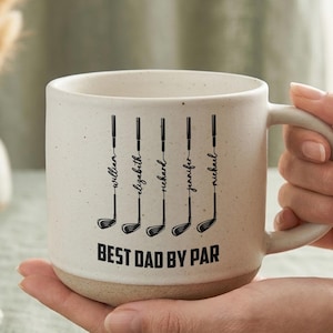 Puede incluir: Una taza de cer&aacute;mica color crema con tem&aacute;tica de golf. La taza presenta ilustraciones de palos de golf negros con nombres encima y la frase "BEST DAD BY PAR" en letras negritas. La taza tiene una textura moteada y una base beige.