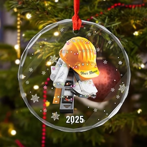 May include: Circular Christmas decoration with a construction theme. It shows a yellow hard hat decorated with fairy lights, architectural plans, and tools. The text "Your Name" and "2025" are displayed on the ornament.