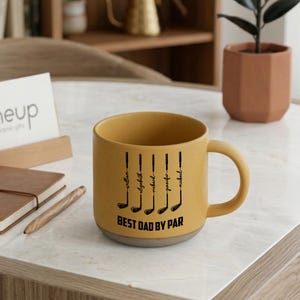 Puede incluir: Taza de cer&aacute;mica amarillo mostaza con base gris. La taza presenta ilustraciones de palos de golf negros y el texto "BEST DAD BY PAR" con nombres. Un gran regalo para los amantes del golf.