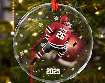 May include: A clear, round Christmas ornament with a hockey player in a red uniform, number 26, and the year 2025. The ornament has a red ribbon for hanging and is decorated with white snowflakes.