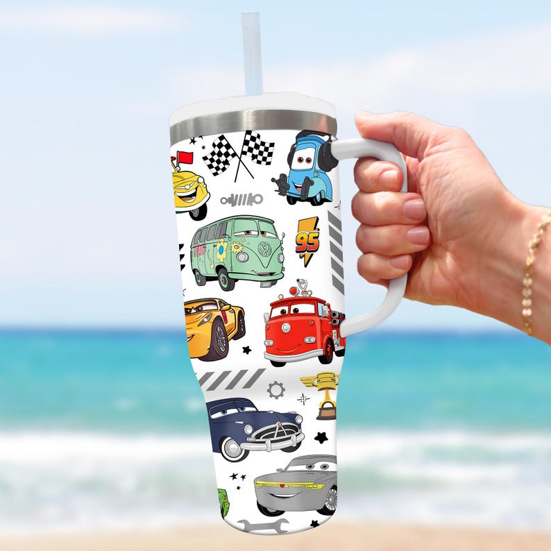 May include: A white tumbler with a handle and a straw, featuring a colorful pattern of cartoon cars. The design includes various car illustrations, checkered flags, and racing-themed elements. The tumbler is held by a person.