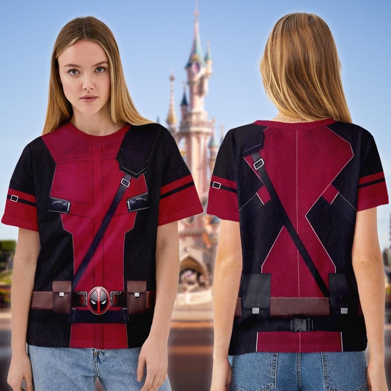 May include: Red and black t-shirt featuring a Deadpool costume design. The shirt has a detailed print of the character's suit, including straps, pouches, and the Deadpool logo. The background shows a castle.