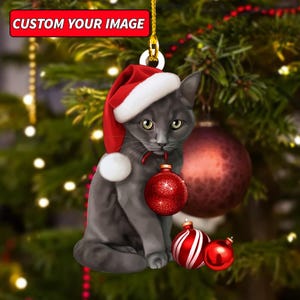 May include: A gray cat ornament wearing a Santa hat and holding a red Christmas ornament. The ornament has the text "CUSTOM YOUR IMAGE" at the top. Additional red and white ornaments are in the foreground, with a Christmas tree in the background.