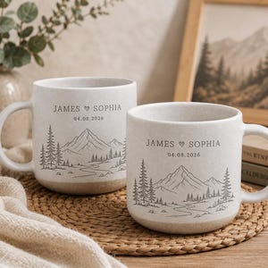 May include: Two off-white ceramic mugs featuring a dark grey line art illustration of a mountain range, pine trees, and a winding river. Each mug is personalised with the text JAMES HEART SOPHIA and the date 04.08.2026 in a simple serif font.