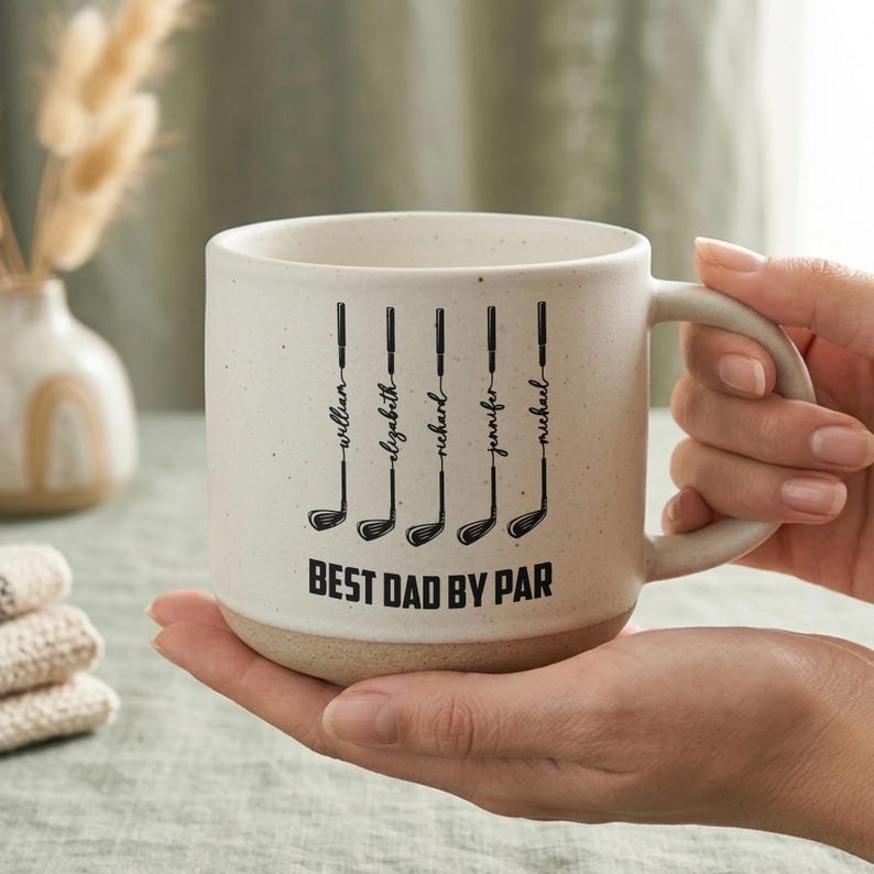 Puede incluir: Una taza de cer&aacute;mica color crema con tem&aacute;tica de golf. La taza presenta ilustraciones de palos de golf negros con nombres encima y la frase "BEST DAD BY PAR" en letras negritas. La taza tiene una textura moteada y una base beige.