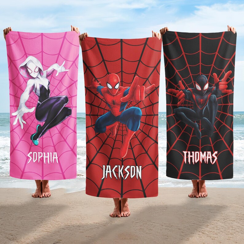 Op de afbeelding: Drie gepersonaliseerde strandlakens met Spider-Man-personages. E&eacute;n is roze met Spider-Gwen, &eacute;&eacute;n is rood met Spider-Man en &eacute;&eacute;n is zwart met Miles Morales. Elke handdoek heeft een naam.
