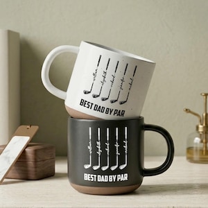 Puede incluir: Dos tazas de cer&aacute;mica apiladas con tem&aacute;tica de golf. La taza superior es blanca con texto negro e ilustraciones de palos de golf, mientras que la taza inferior es negra. Ambas tazas dicen "BEST DAD BY PAR" y tienen nombres personalizados.