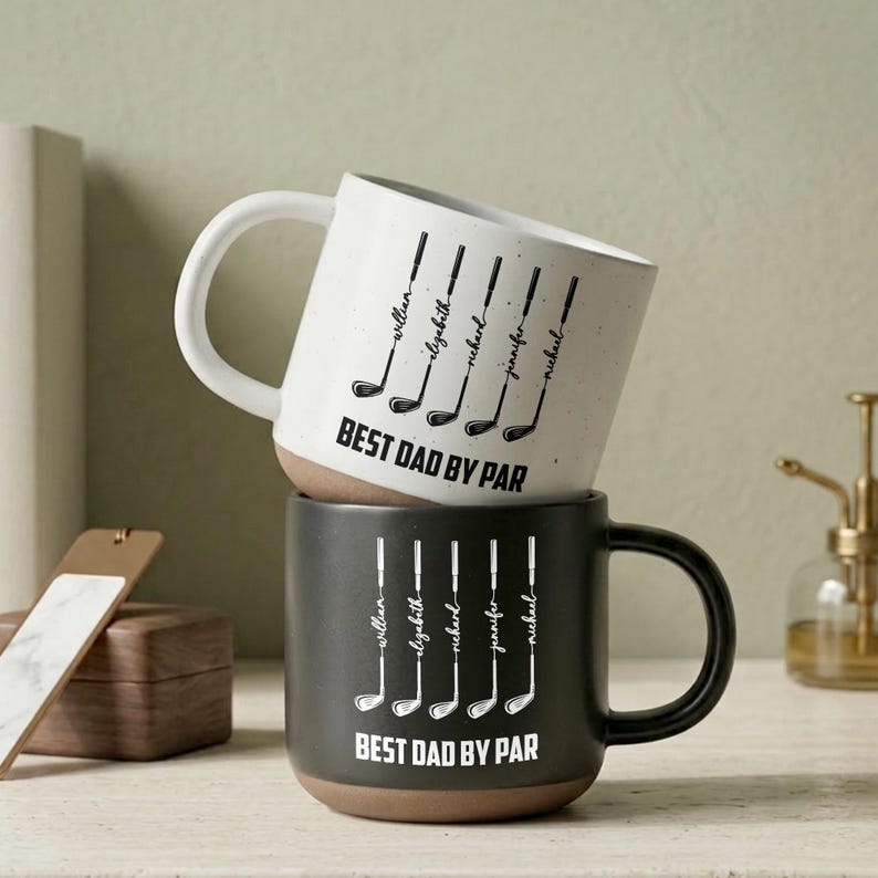 Puede incluir: Dos tazas de cer&aacute;mica apiladas con tem&aacute;tica de golf. La taza superior es blanca con texto negro e ilustraciones de palos de golf, mientras que la taza inferior es negra. Ambas tazas dicen "BEST DAD BY PAR" y tienen nombres personalizados.