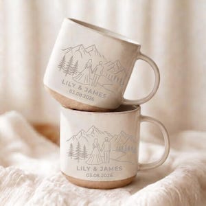 May include: Two off-white ceramic mugs stacked vertically, each featuring a debossed line art illustration of a couple standing before mountains and pine trees. The text LILY & JAMES and the date 03.08.2026 are engraved below the illustration.