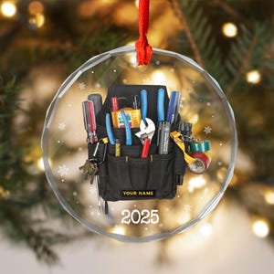 May include: A clear, round Christmas ornament with a black tool belt design. The tool belt is filled with various tools, including screwdrivers, pliers, and a wrench. The text "YOUR NAME" and "2025" are displayed on the ornament. A red ribbon is attached for hanging.