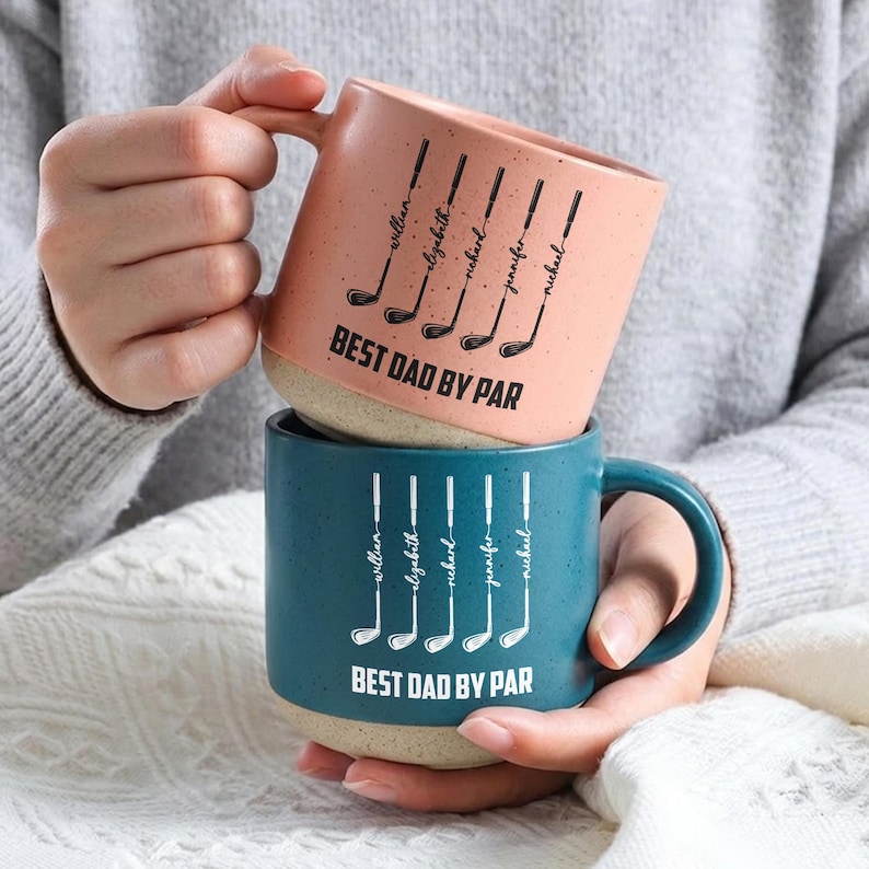 Puede incluir: Dos tazas de cer&aacute;mica, una coral y otra turquesa, apiladas. Cada taza presenta un dise&ntilde;o de palo de golf con nombres y el texto "BEST DAD BY PAR". Las tazas tienen un acabado moteado y una base beige.