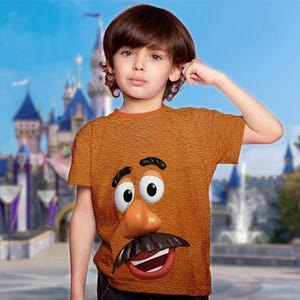 May include: A brown t-shirt featuring a large, cartoonish face with a mustache, nose, and eyes. The shirt has a textured appearance and is worn by a child. The background shows a castle and blue sky.
