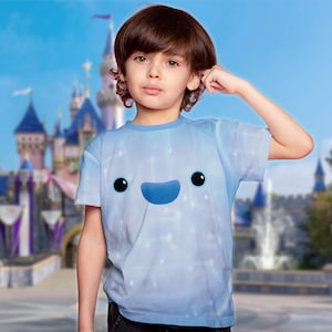 May include: A light blue t-shirt featuring a cartoon face with black eyes and a blue smile. The shirt has a gradient effect with white dots. The background shows a castle and blue sky.