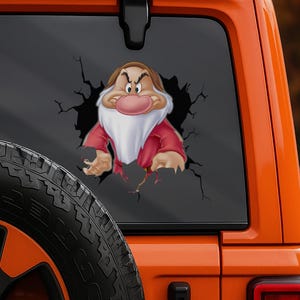 May include: A decal featuring Grumpy from Snow White, bursting through a cracked black background. Grumpy is depicted with a red coat, white beard, and a grumpy expression. The decal is applied to the rear window of an orange vehicle.