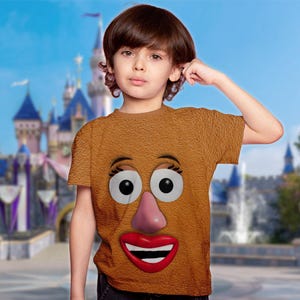 May include: A brown t-shirt featuring a cartoon face with large white eyes, a pink nose, and a red smiling mouth. The shirt is worn by a child in front of a blurred castle background.