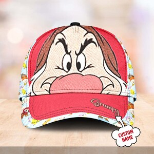 May include: Red baseball cap with a colourful pattern featuring the character Grumpy from Snow White. The front of the cap features a close-up of Grumpy's face with the text "Grumpy" printed below.