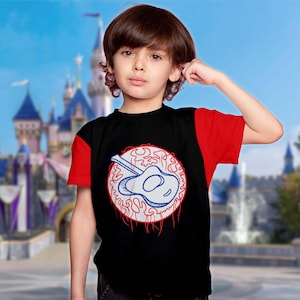 May include: Black and red short-sleeved t-shirt with a white and blue guitar design within a red and white circle. A young person with brown hair is wearing the shirt, with a blurred castle in the background.